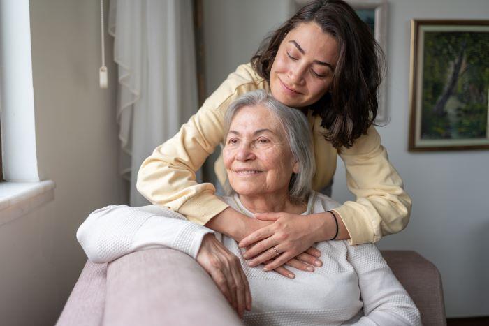 Mother seated and adult daughter standing behind her with her arms around her mother