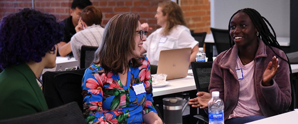 Three students have a dynamic conversation in a workgroup, while their fellow classmates hold separate discussions in the background