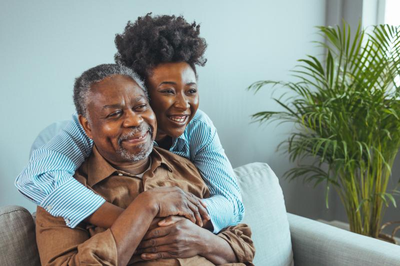 A daughter embraces her elderly father from behind. Both are smiling.