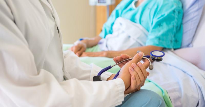 A close up image of a doctor's hands holding a stethoscope while having a discussion with a patient sitting in a hospital bed.