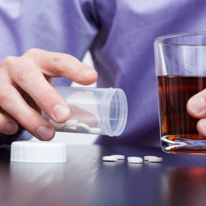 A man dispenses pills on a tabletop while holding an alcoholic beverage in his other hand.