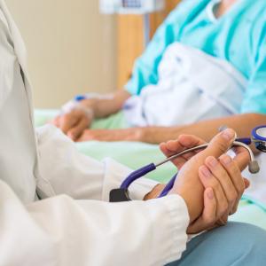 A close up image of a doctor's hands holding a stethoscope while having a discussion with a patient sitting in a hospital bed.