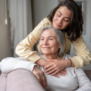 Mother seated and adult daughter standing behind her with her arms around her mother
